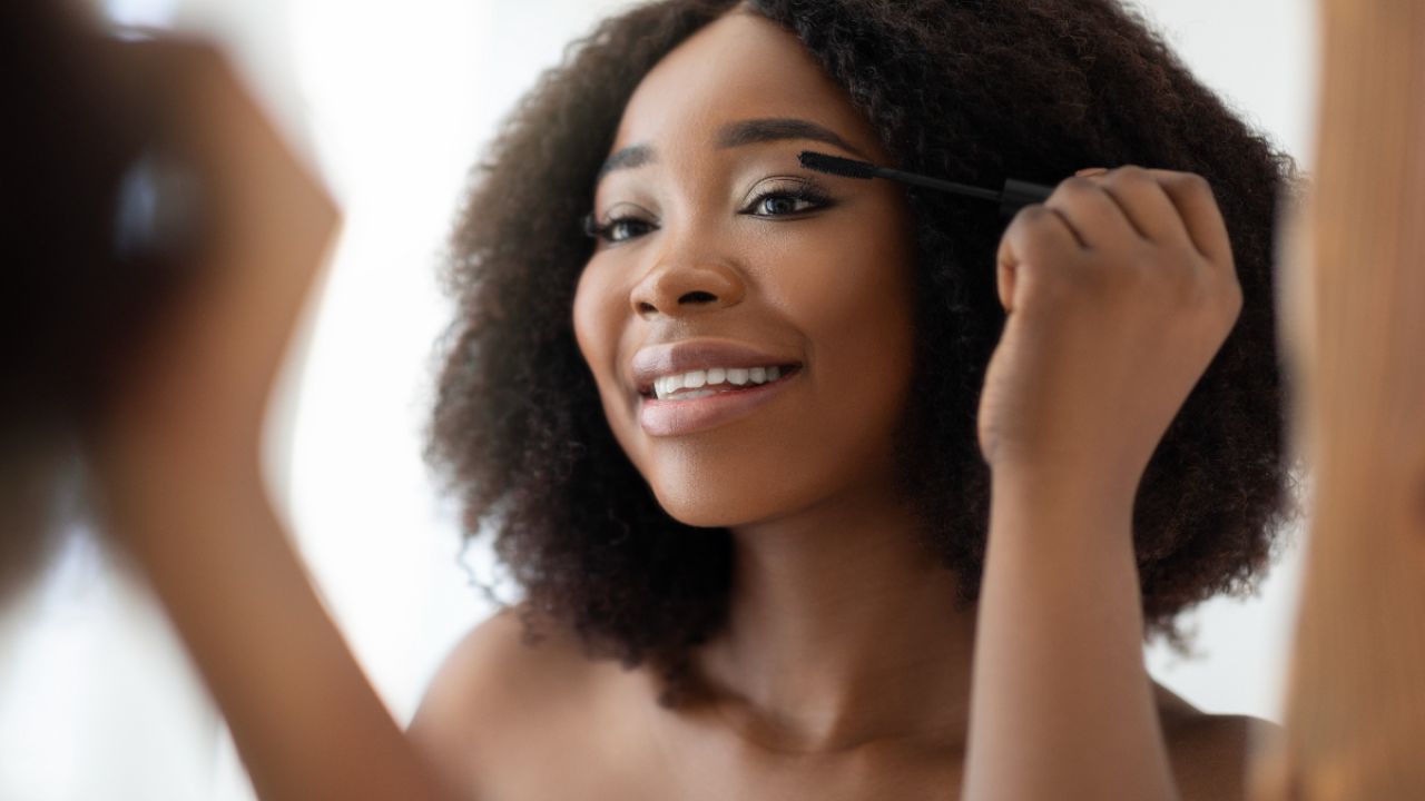 A woman with curly hair applies mascara while smiling in front of a mirror.