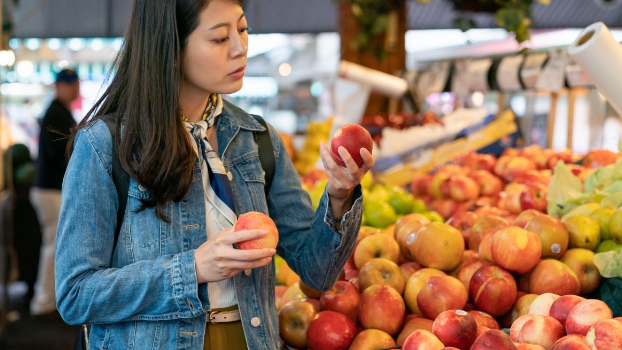 Photo: woman in denim jacket examining a red apple at a busy market stall