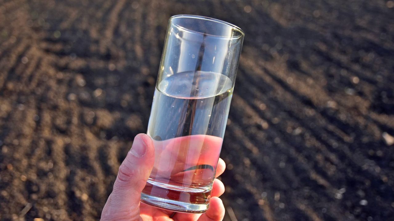 Photograph of hand holding clear glass of water above plowed field