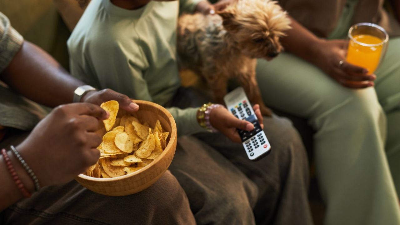Photo: family sharing chips on couch with small dog, TV remote and glass of orange juice