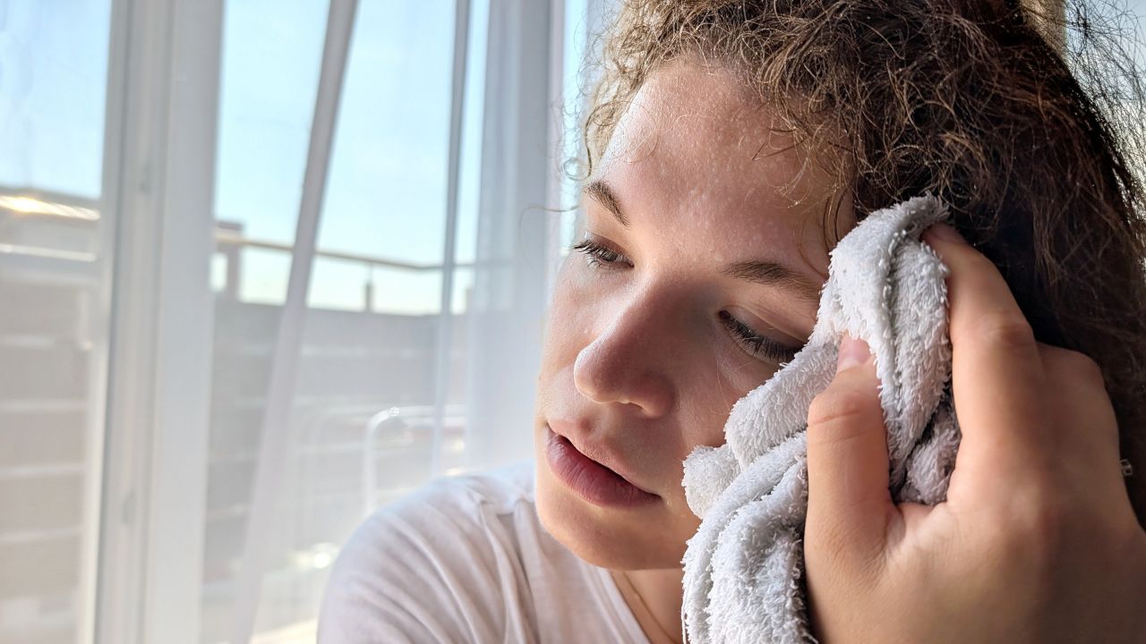 Photograph of a young woman with eyes closed pressing a towel to her cheek by sunlit curtains.